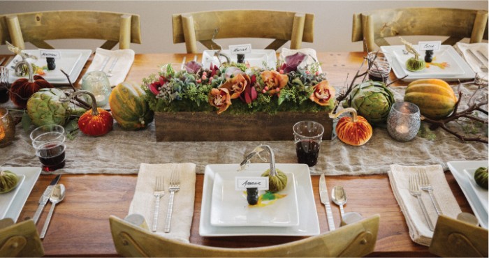 thanksgiving centerpiece, small and narrow wooden crate, filled with green plants, and orange flowers, surrounded by pumpkins, gourds and dried branches, on a wooden table with square, white plates