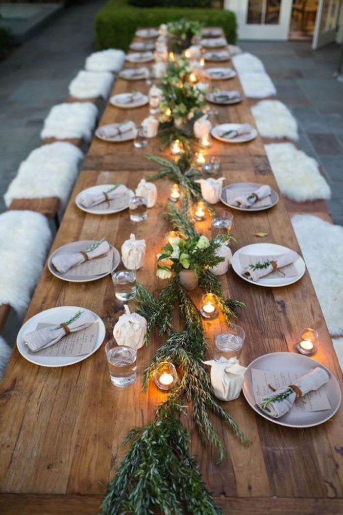 eighteen plates with napkins, placed on a long, rustic wooden table, decorated with green branches, and tea lights