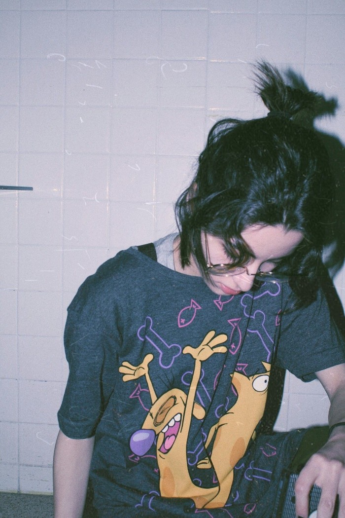 young woman with dark hair, partially styled in a messy half-bun, wearing a dark grey t-shirt, with a colorful 90s grunge cartoon print
