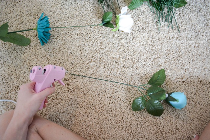 glue gun in pink, held by a pale hand, near a faux blue rose, with green plastic leaves, and a long stalk, diys for your room, more fake flowers nearby