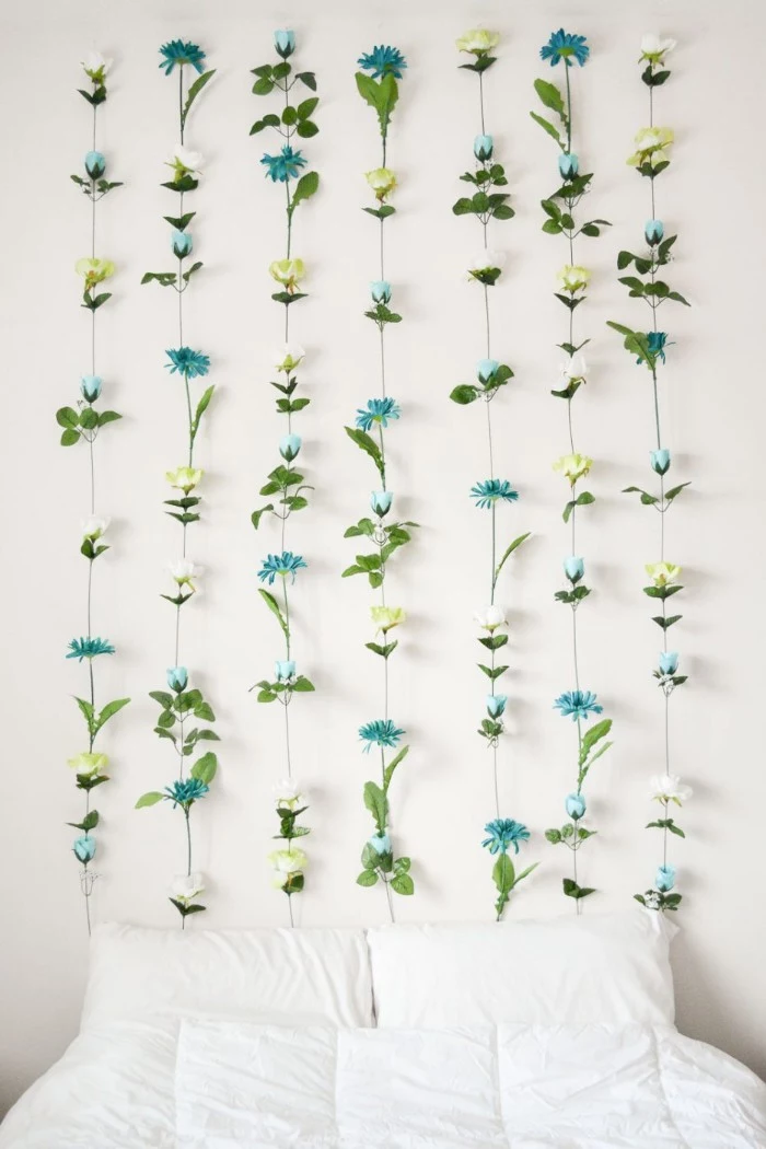 blue and light yellow, and white faux flowers, with green leaves, and long plastic stems, glued together in seven columns, and attached to a white wall, near a bed