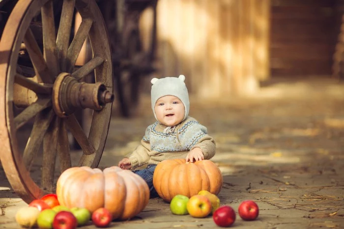 pumpkins and apples, near a sitting baby boy, dressed in a beige and light blue sweater, and dark blue jeans, baby thanksgiving outfits, pale grey hat, with small bear ears