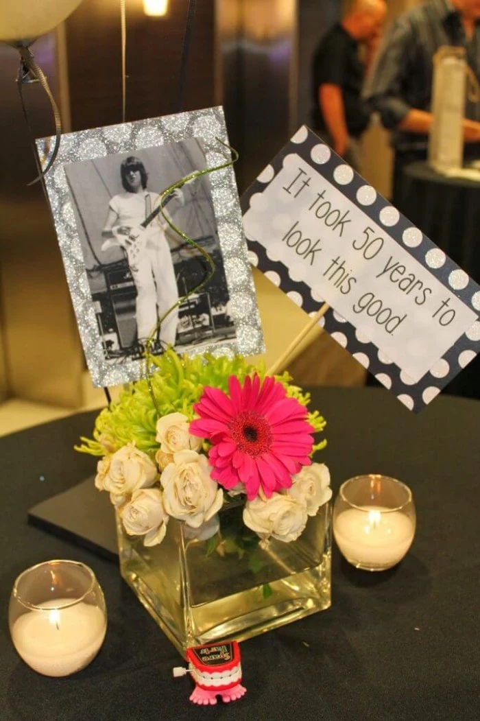 cube-shaped vase, made from clear glass, and containing a small bouquet, decorated with a black and white photo of a young man, and a sign reading, it took 50 years to look this good