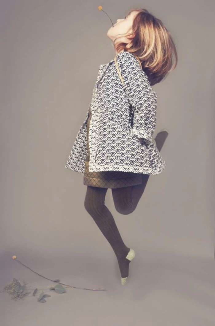 little girl with shoulder-length blonde hair, and a yellow flower in her mouth, jumping in the air, dressed in a white and black patterned coat, a khaki short dress, and dark grey knitted tights, girls thanksgiving outfit