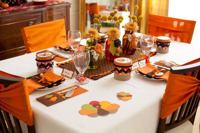 children's thanksgiving table, white tablecloth and orange napkins, brown plates with white pollka dots, three vases in different colors, containing yellow flowers, cheap centerpiece ideas, multicolored turkey shaped paper cutouts