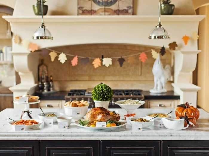 paper garland featuring fall leaves, in different colors, hanging near an antique-style stove, thanksgiving wishes, kitchen island covered in different dishes and foods