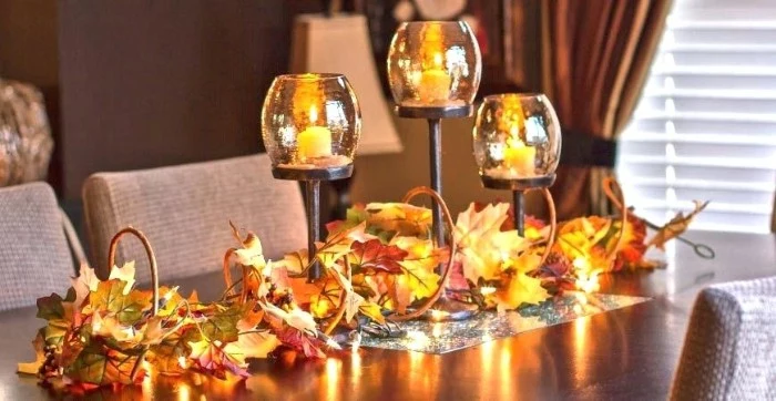 candle holders made of glass and metal, each containing a lit, small yellow candle, on a dark brown table, decorated with orange and yellow, faux fall leaves
