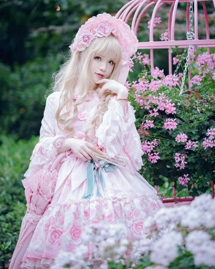 garden with pink blossoms, behind a girl in a blonde wig, wearing a white frilly dress, decorated with a subtle rose pattern, she holds a fan and a pink parasol