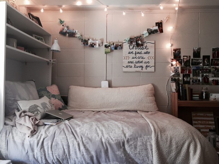 lit string lights, hanging over a bed, with light cream, and pale grey, covers and pillows, in a dim room, with shelves and wall decorations