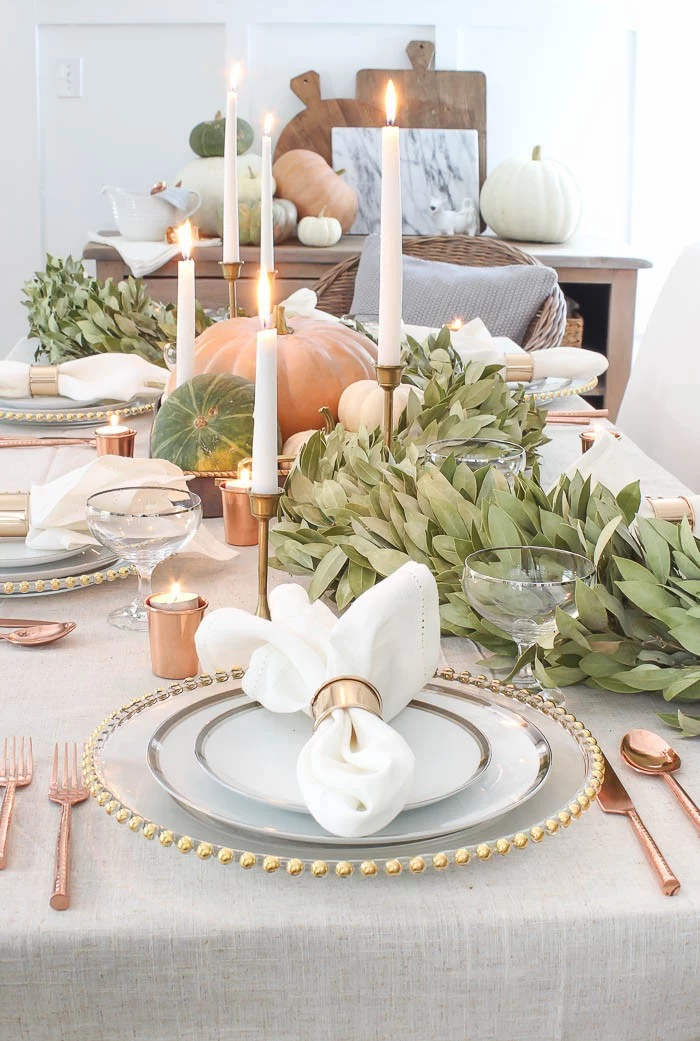 rose gold cutlery, placed near three stacked plates, with gold and silver details, thanksgiving table decorations, lit candles and several pumpkins, green decorative plants