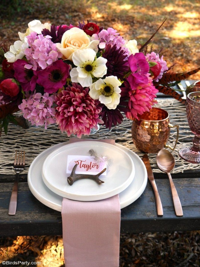 copper mug and two stacked white plates, topped with a small dark brown antler, and a name card, pale pink napkin, and some cutlery, on a table, decorated with a large bouquet 