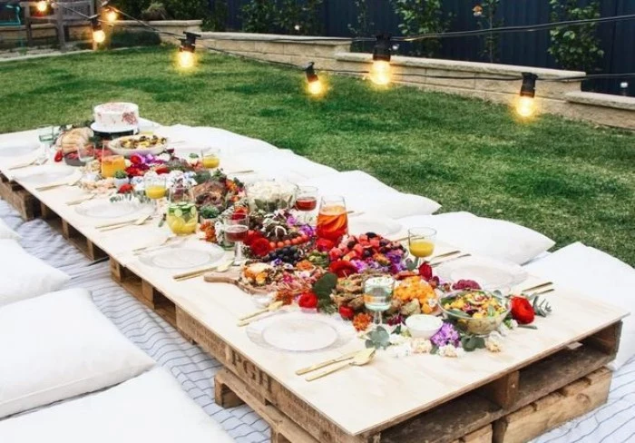 pallets used to make a table, set up for a festive meal, and decorated with a variety of flowers, 50th birthday party ideas for mom, boho garden picnic, with string lights overhead