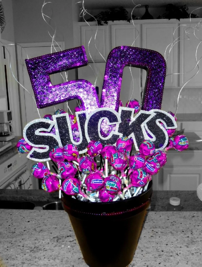 glittering purple decoration, reading 50 sucks, placed in a small bucket, filled with lolipops, in purple wrappers