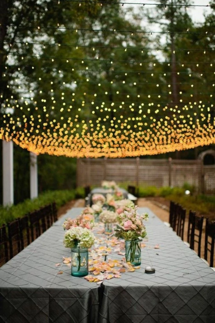 50th birthday party ideas for mom, long table decorated with flower petals, and several small bouquets in mason jars, inside a garden, mesh of fairy lights overhead