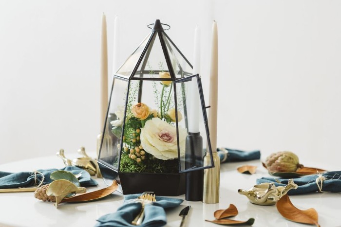 terrarium made of glass, with black frames, containing a white flower, and several yellow blossoms, thanksgiving table decorations, on a round white table, sprinkled with fall leaves