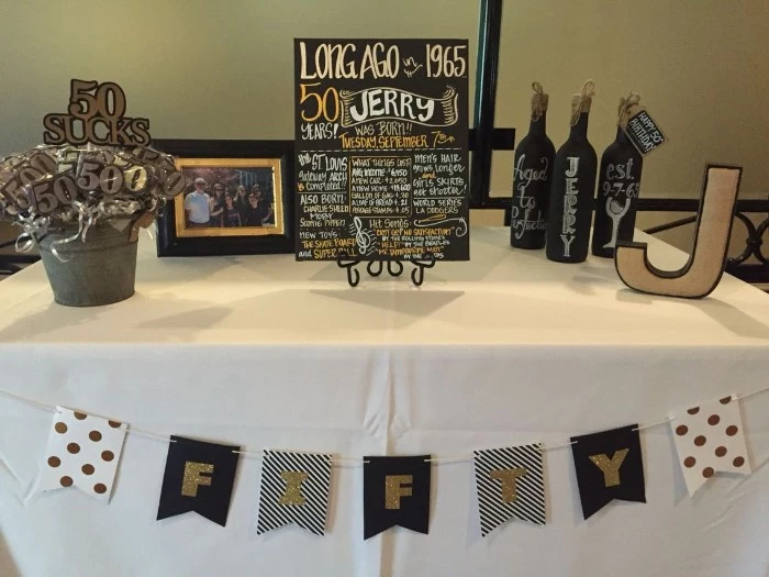 simple birthday decor, table with a festive poster, three hand-painted wine bottles, a bucket with lollies shaped like the number 50, a j-shaped decoration, and a family photo in a frame