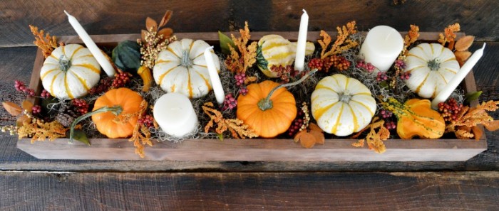 six white candles, in different shapes, placed in a narrow wooden crate, containing white and orange pumpkins, gourds and fall leaves, thanksgiving centerpiece, on a dark brown wooden table