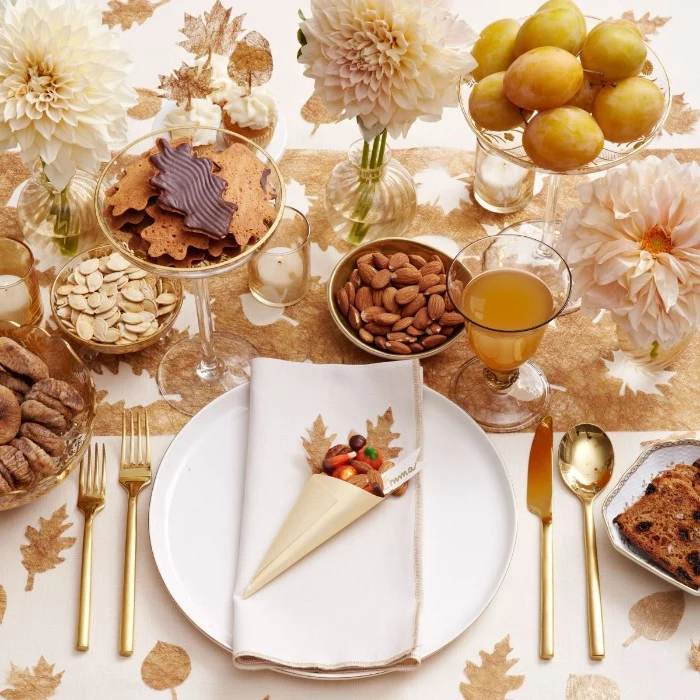 bisquits and fruit, nuts and seeds, in various dishes, placed on a white tablecloth, decorated with pale beige leaves, thanksgiving table setting, white plate and napkin, gold cutlery and a glass with juice