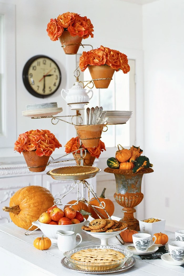 ceramic pots filled with orange flowers, placed on a metal stand, with a pie, some cutlery and plaates, and a white pot, thanksgiving table decorations