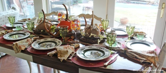 set table with eight plates, eight clear green wine glasses, floral napkins and silver cutlery, and a centerpiece, featuring several wrapped pumpkins, near a several windows