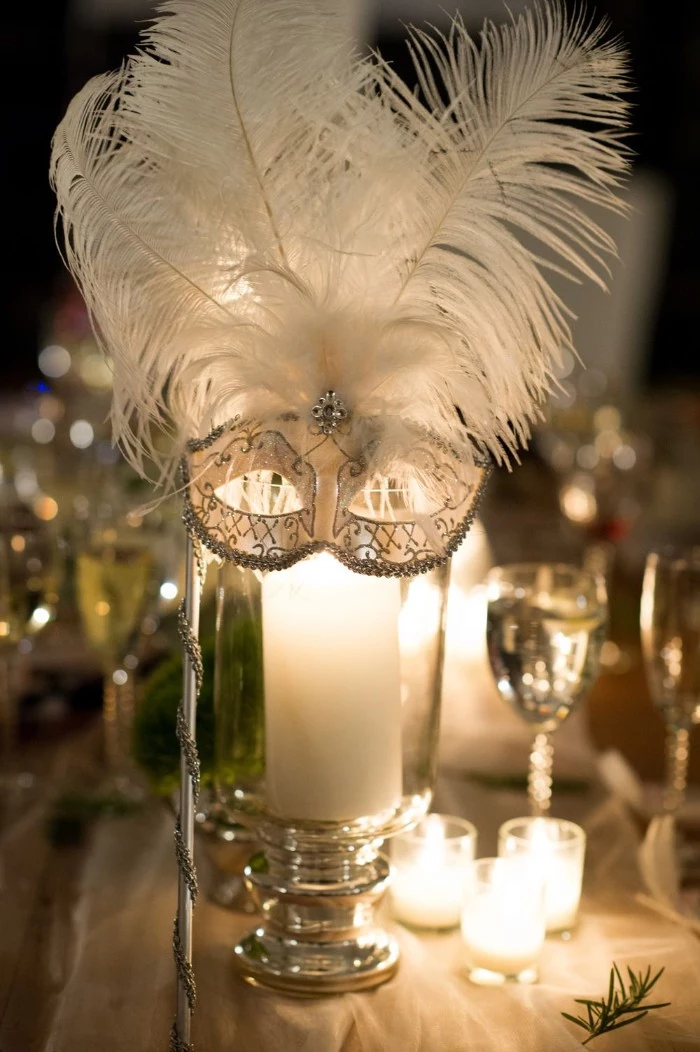 venetian-style mask in white, decorated with silver glitter, and large white ostrich feathers, placed on a glass container, with a large lit candle, 50th birthday ideas, fancy masked ball