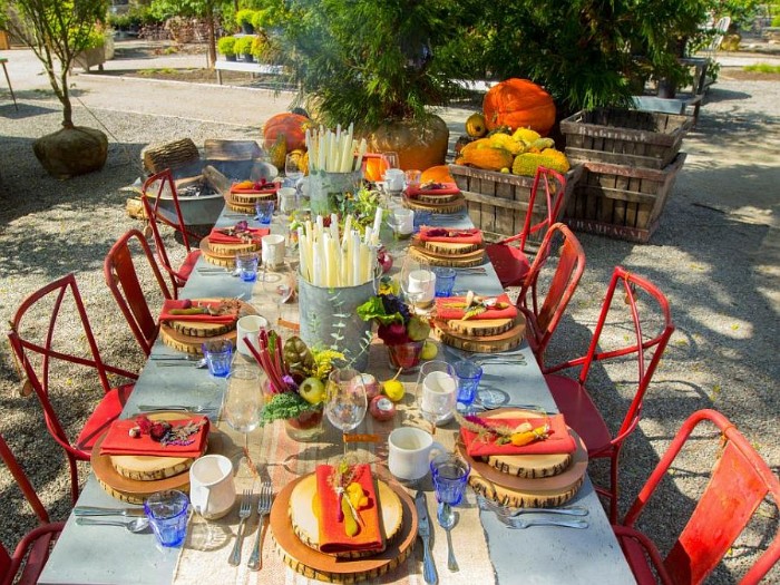 red chairs near a pale blue, rectangular dinner table, set for eight people, and placed outdoors, decorated with two buckets, filled with candles, and bowls of fall vegetables