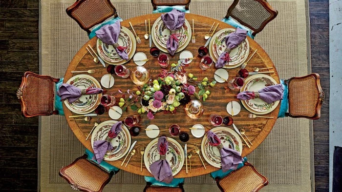 areal shot of an oval wooden table, with eight chairs, and eight ornamental plates, with folded violet napkins, large bouquet in the center of the table