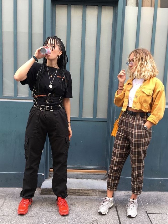 two young women, dressed in 90s grunge fashion, one wearing a black outfit, with multiple belts and red sneakers, the other in checkered ankle trousers, white cropped top, and a cropped yellow jacket