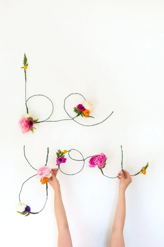 hands holding the word you, made from thin, green wire, and decorated with faux flowers, in different colors, diys for girls, the word be, created in a similar way, is mounted on the wall above