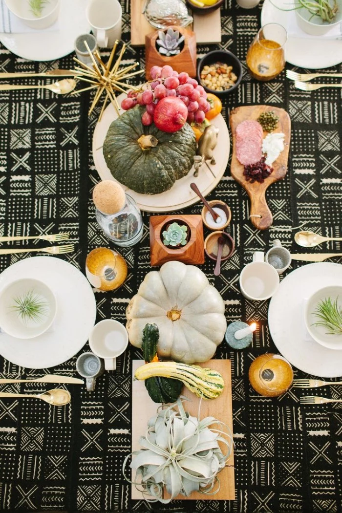 pomegranates and grapes, pumpkins and gourds, decorating a thanksgiving table, covered with a black and white, patterned tablecloth with plates, cutlery and glasses