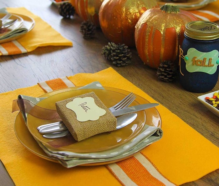 close up of two stacked, dark yellow plates, with a pale grey napkin between them, cutlery and a small square object, wrapped in brulap and decorated with a name tag, placed on top, thanksgiving tablescape