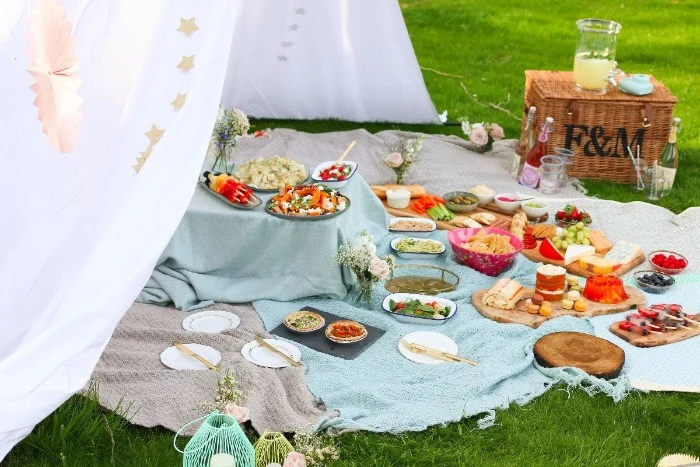 50th birthday ideas, green lawn with several blankets, covered in various dishes, large white fabric tent-like structure, picnic basket and decorations