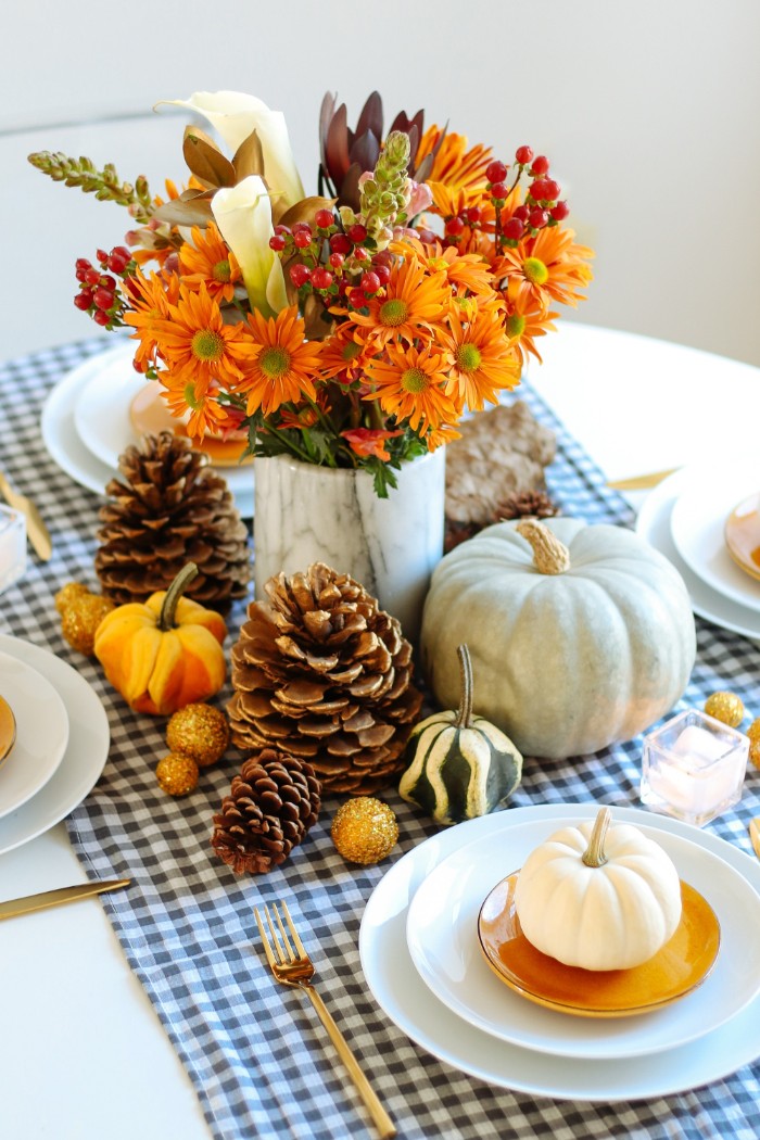 bouquet with orange, white and red flowers, in a marble vase, surrounded by pumpkins in different sizes, pinecones and yellow baubles, thanksgiving table decorations, plates and cutlery