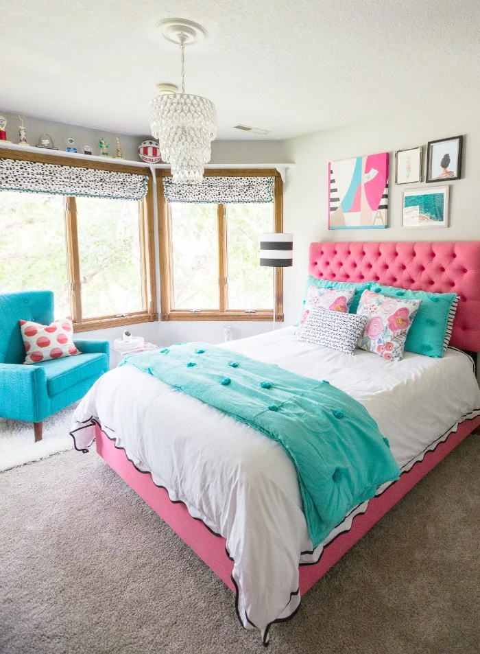 soft headboard in pink, on a pink bed, with white and teal covers, and five cushions, in teal and white and multicolor, two large windows, and wall decorations