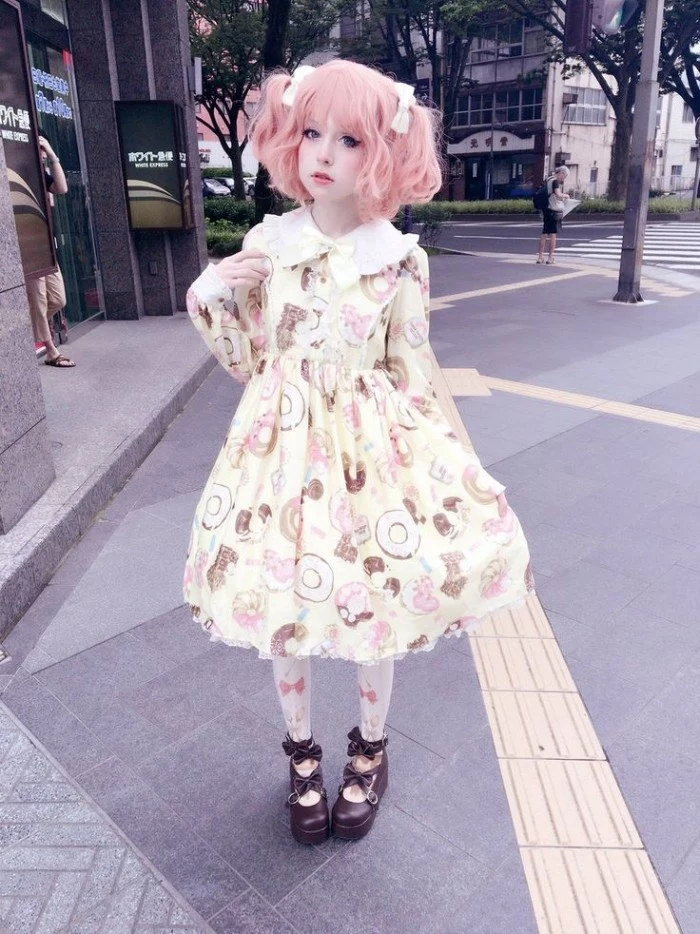 short pink wig, featuring pigtails decorated with white bows, on a pale girl, dressed in a cream dress, with a frilly white collar, and a pattern with colorful donuts and sweets