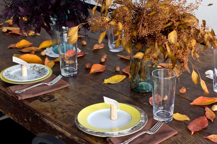 vintage dark brown wooden table, with yellow and white plates, each containing a small name card, propped up on a cork bottle stopper, bouquets of dried plants, yellow and orange, red and brown leaves strewn about the table