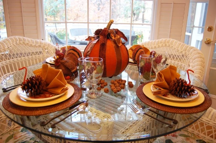 dishes decorated with turkey ornaments, made from pinecones, orange pipe cleaners, and yellow napkins, folded like fans, on a round glass table, near a large pumpkin, and a turkey figurine