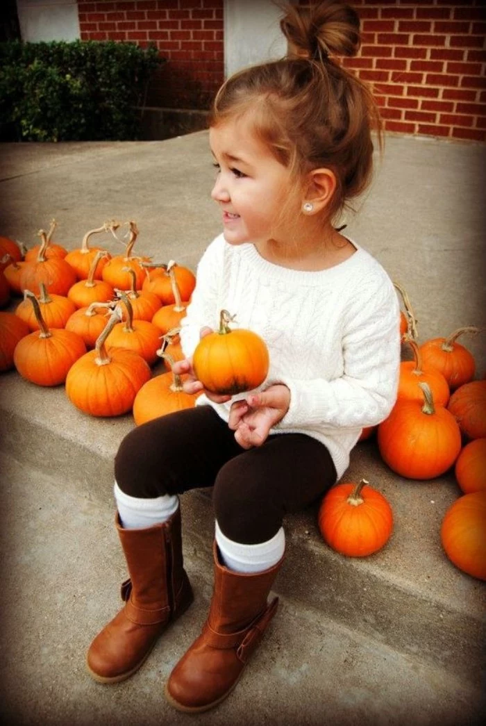 child dressed in a white cable knit jumper, black leggings with white socks, and tall brown leather boots, smiling while holding a small pumpkin, toddler thanksgiving outfit, more pumpkins nearby