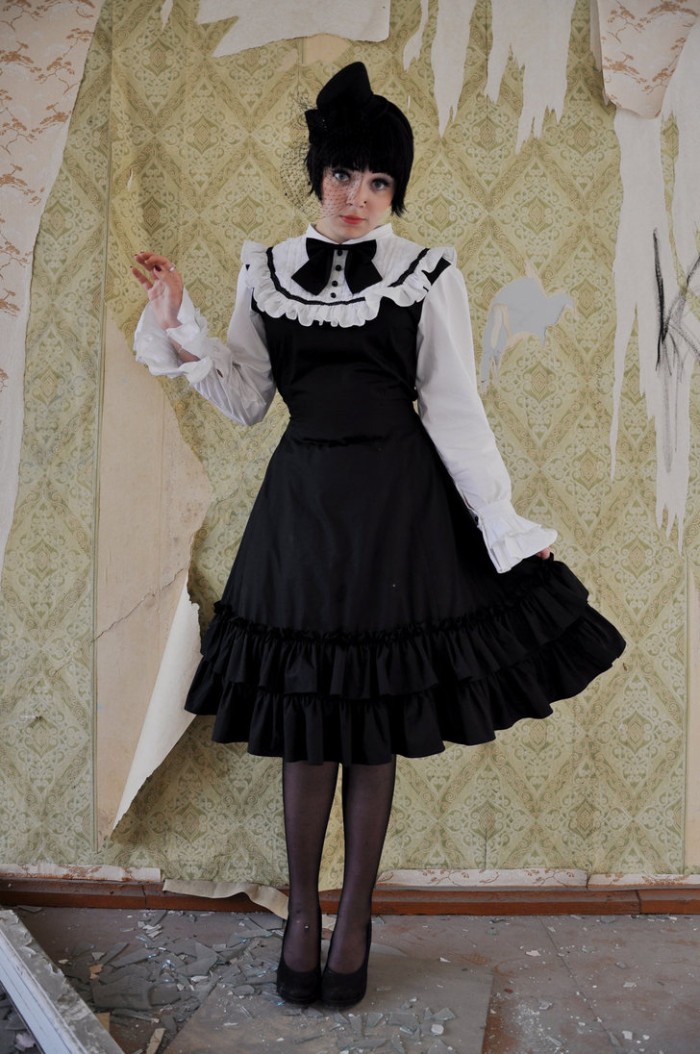 old ripped ornamental wallpaper, behind a young woman, in a black wig, wearing a black and white dress, with frills and a bow, define lolita