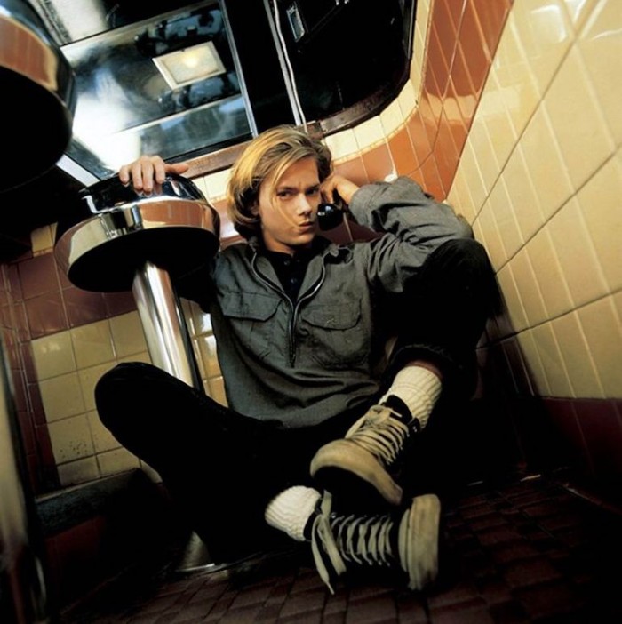 actor river phoenix, sitting on the floor of a retro diner, holding a telephone receiver, and leaning on a bar stool, 90s bands and iconic celebrities