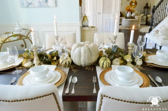dining room with a dark brown wooden table, decorated with pumpkins, in different sizes and colors, four lit white candles, plates and bowls, napkins and cutlery