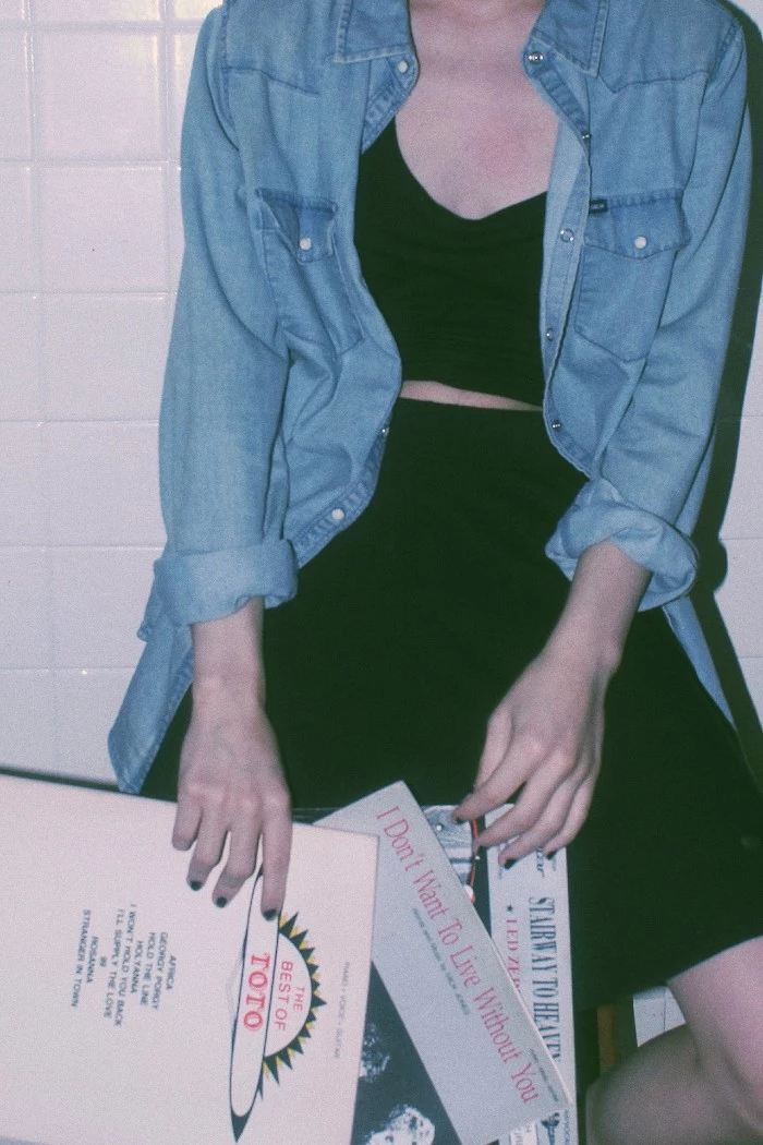 chambray shirt in light blue, worn unbuttoned over a black mini skirt, and a black cropped top, by a pale woman, holding three record cases