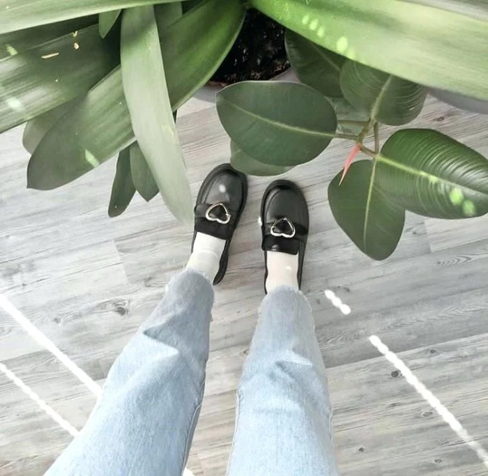 house plants with different, large green leaves, near a pair of legs, dressed in pale blue, acid wash cutoff ankle jeans, worn with white socks, and black loafers, featuring silver heart-shaped buckles, grunge girl