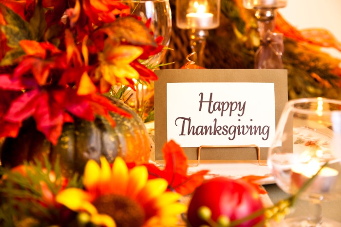 card with the words happy thanksgiving, placed on a table, decorated with multiple faux flowers, in red and yellow and orange, thanksgiving message to employees
