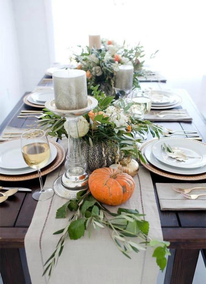thanksgiving tablescape, dark wooden table, decorated with small orange pumpkins, green leaves and silver candles, on clear glass holders, white and gold plates, one wine glass and silver cutlery
