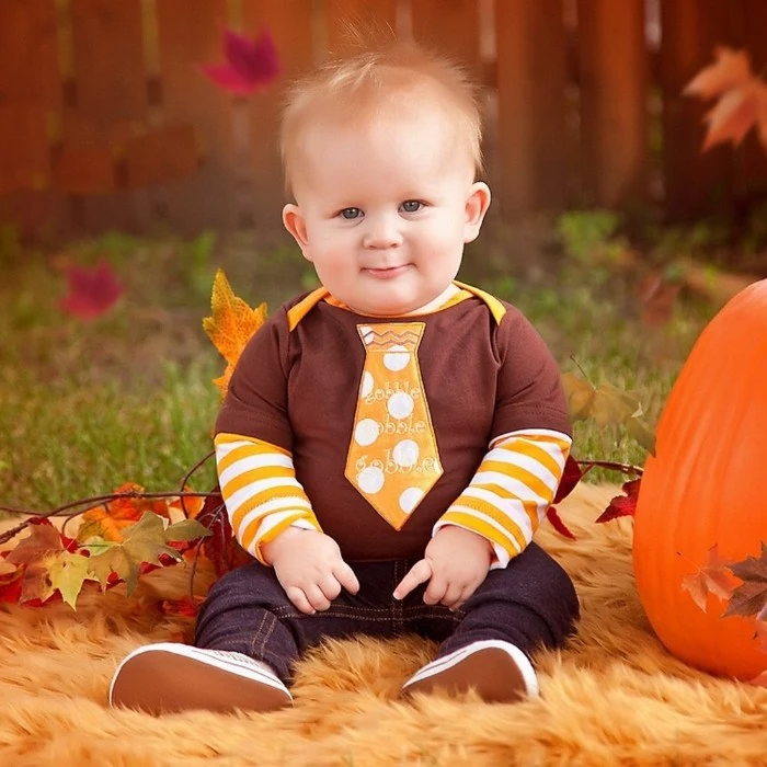 necktie shape in orange, with white polka dots, sewn on a brown t-shirt, worn over a white and orange striped jumper, by a smiling baby boy, baby's first thanksgiving outfit, dark blue jeans