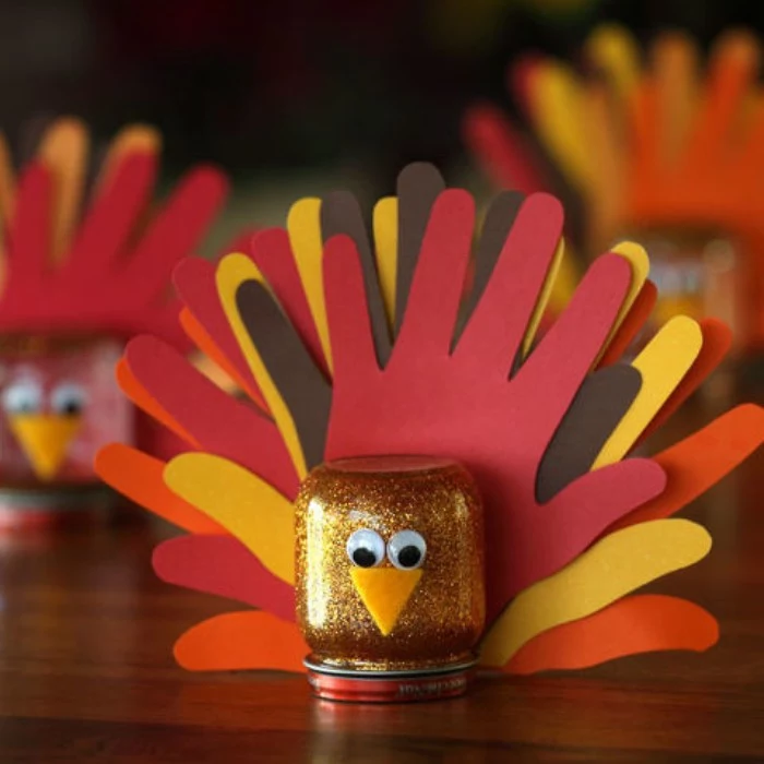 kids diy turkey decorations, small mason jar filled with gold glitter, decorated with eye stickers, a small felt beak, and a tail, made from hand-shaped paper cutouts in different colors