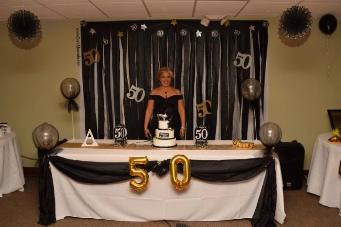 blonde woman in a black dress, smiling while standing next to a festively decorated table, 50th birthday party ideas for mom, balloons and a cake