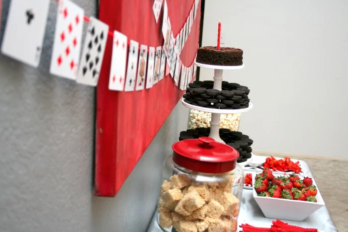 strawberries and cookies, cakes and popocorn, in dishes and glass containers, placed on a table, near a wall, decorated with diy playing card garlands, 50th birthday party ideas for men, easy and cheap