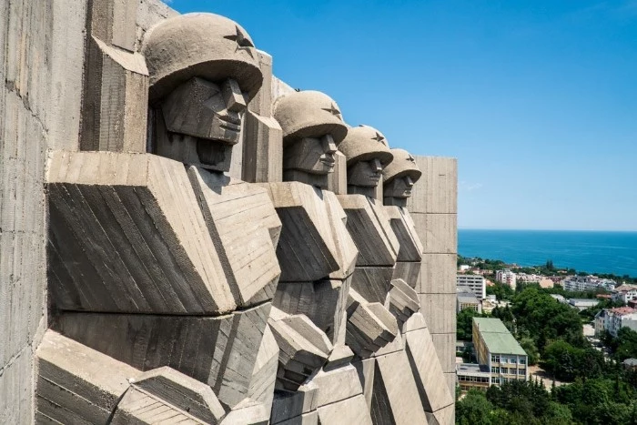 brutalist design of a monument, featuring four soldiers, with helmets decorated with stars, a city near the sea, visible in the background